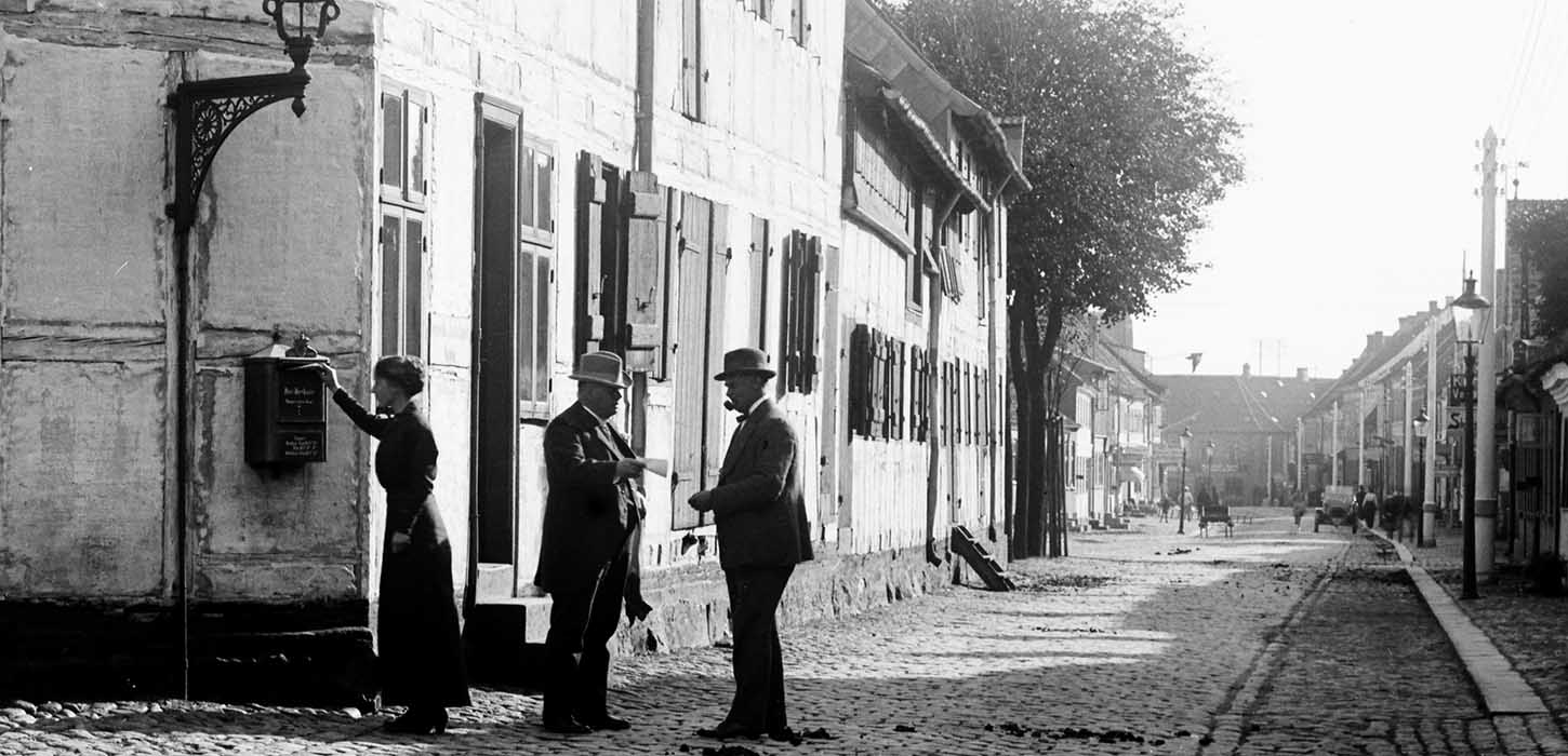 En kvinde står ved siden, og to mænd med bowlerhatte på taler sammen ved Trægården i gamle dage